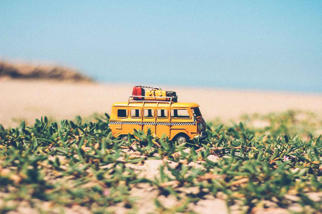 Fotografía artística de una miniatura de furgoneta Volkswagen amarilla con equipaje en el techo, situada sobre una vegetación baja y delante de un fondo desenfocado que sugiere una playa arenosa. La imagen evoca un sentido de aventura y viaje.
