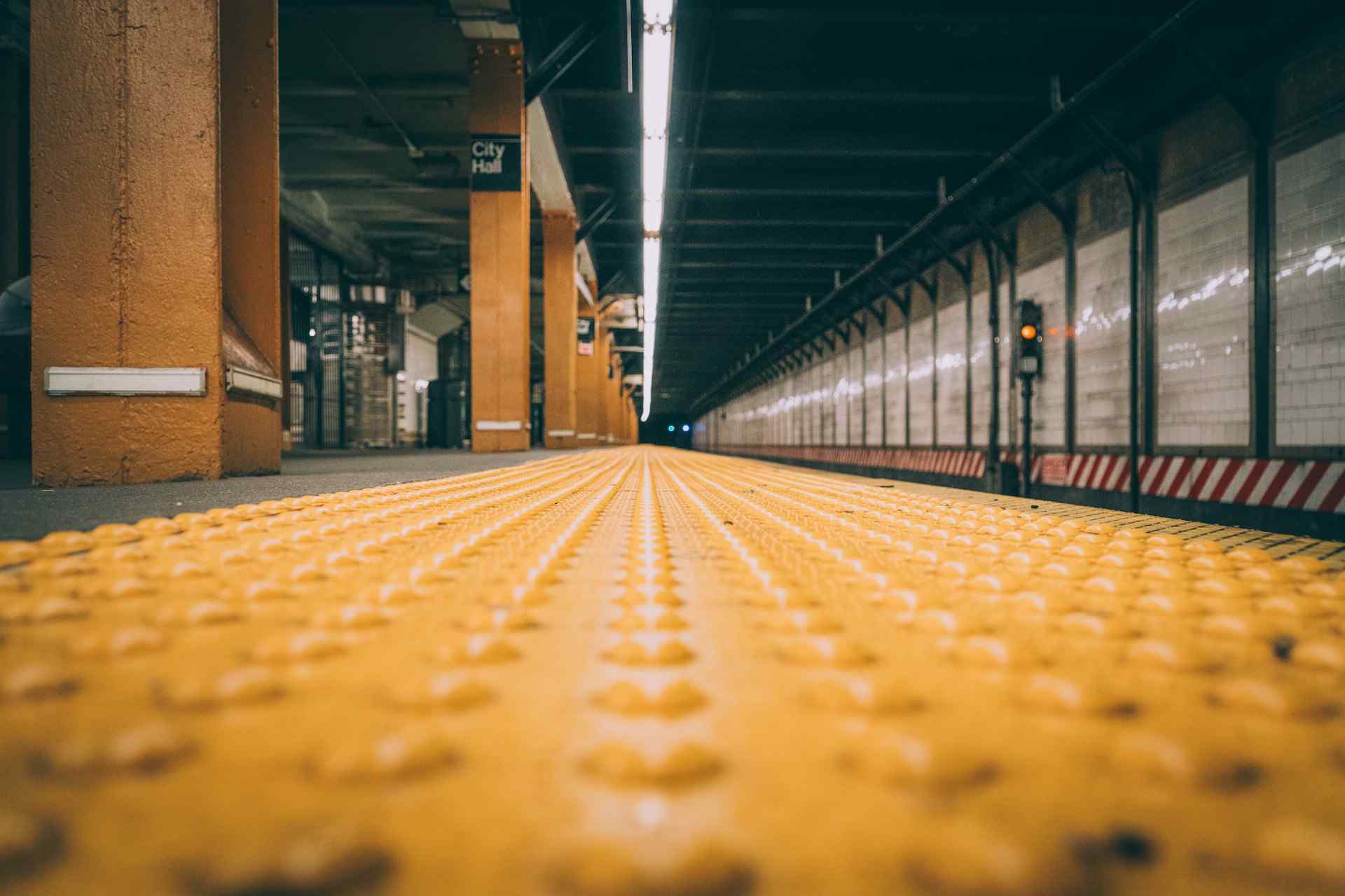 Vista a nivel de suelo de una plataforma de estación de metro con superficie táctil amarilla de advertencia y columnas color naranja, con señalización de 'City Hall' al fondo, enfatizando la accesibilidad y orientación en el transporte público.