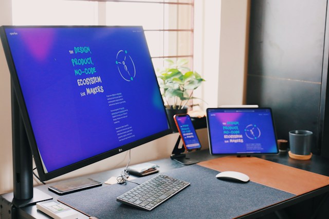 Espacio de trabajo tecnológico moderno con un gran monitor, una laptop, un teléfono inteligente y una tablet alineados en un escritorio, todos con pantallas que muestran una interfaz de usuario uniforme, reflejando un entorno de diseño y desarrollo sin código.