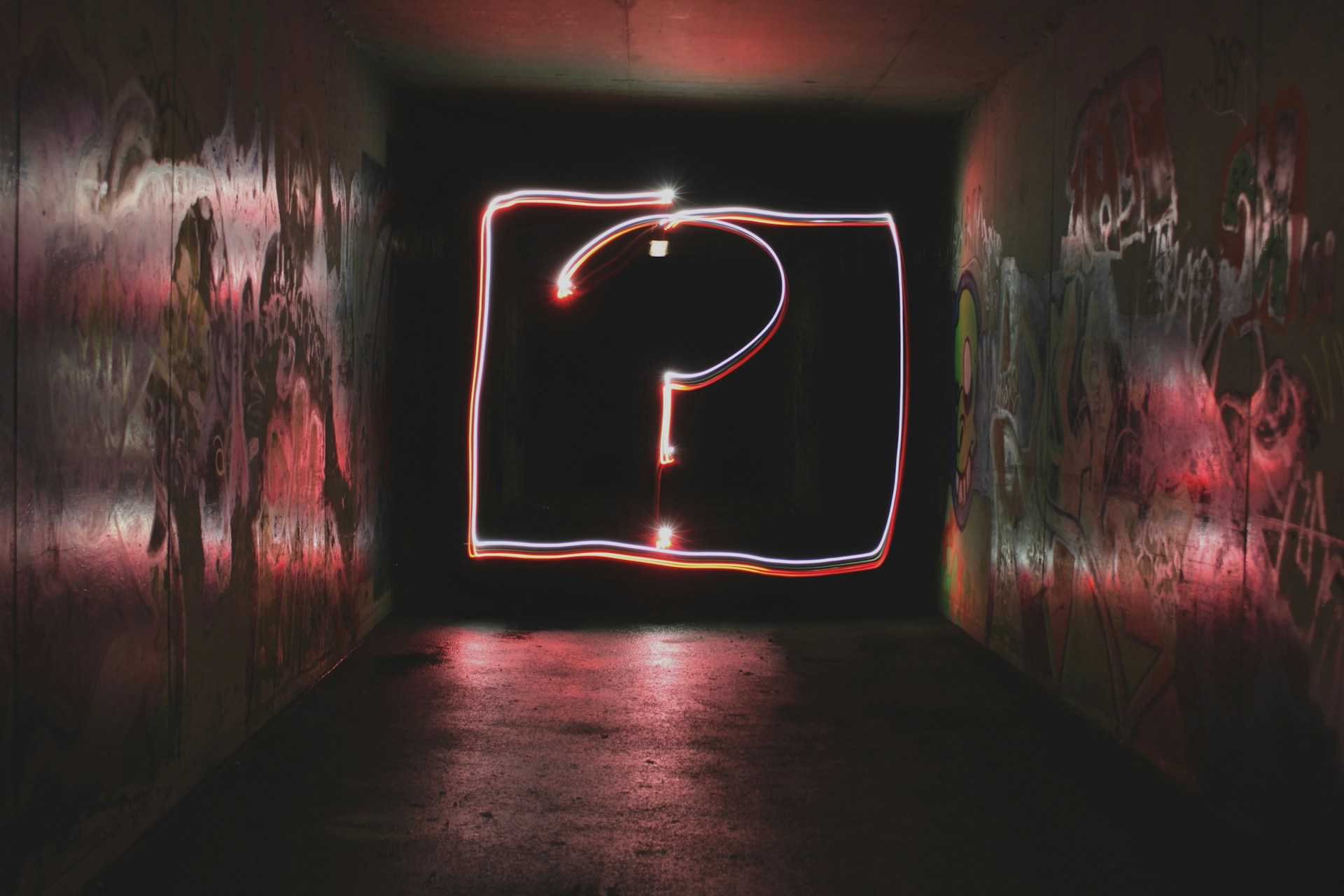 Un gran signo de interrogación de neón rojo y blanco ilumina una pared grafitada en un túnel oscuro, creando un ambiente de misterio y curiosidad.