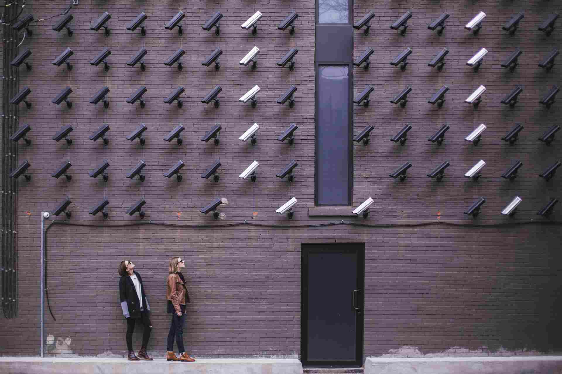 Dos personas de pie junto a una pared de ladrillos oscuros observan numerosas cámaras de seguridad apuntando en su dirección. La escena da una sensación de vigilancia.