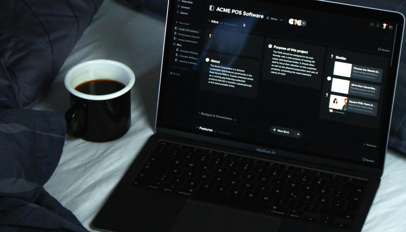 Un MacBook Air mostrando una interfaz de usuario con diseño de modo oscuro para software de punto de venta (POS), con un texto visible que detalla características y propósito del proyecto, acompañado por una taza de café negro en una superficie de cama con sábanas grises.
